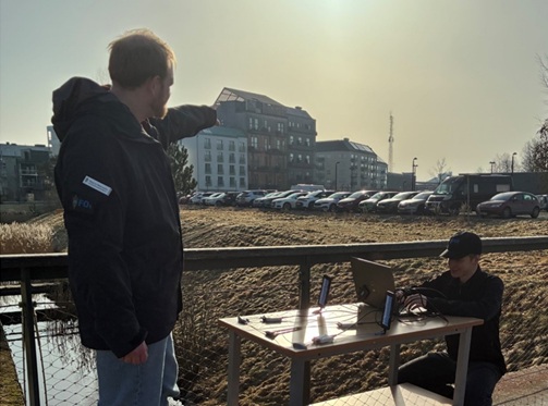 En forskare sitter framför ett bord med en dator och olika typer av teknisk utrustning framför sig. Brevid står en annan forskare, som pekar mot byggnader en bit bort. Personerna sitter utomhus.